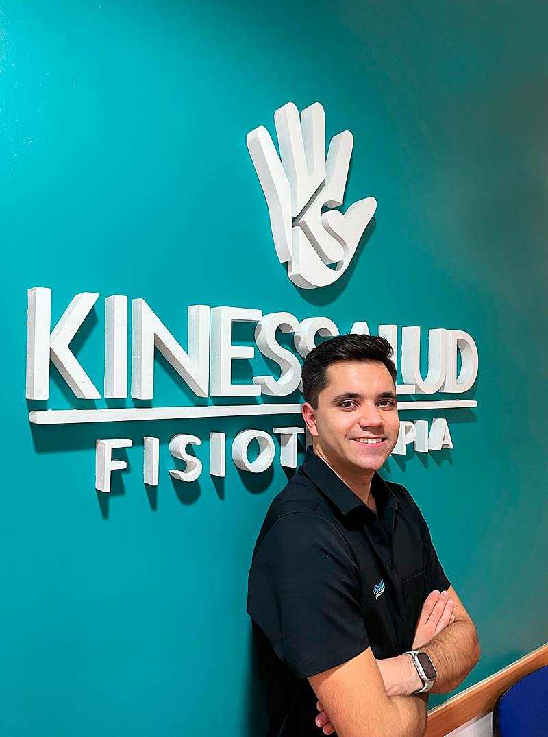 Hombre sonriente posando frente a una pared que dice Kinessalud Fisioterapia.